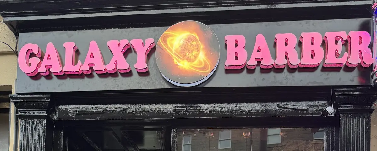 Galaxy Barber's hot pink neon sign glowing after dark, with orange ringed-planet logo between the words